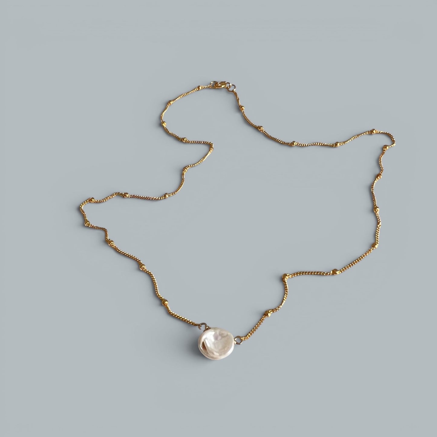 TSJ gold-tone chain necklace with a single, irregular white baroque pearl pendant. Minimalist, elegant design.