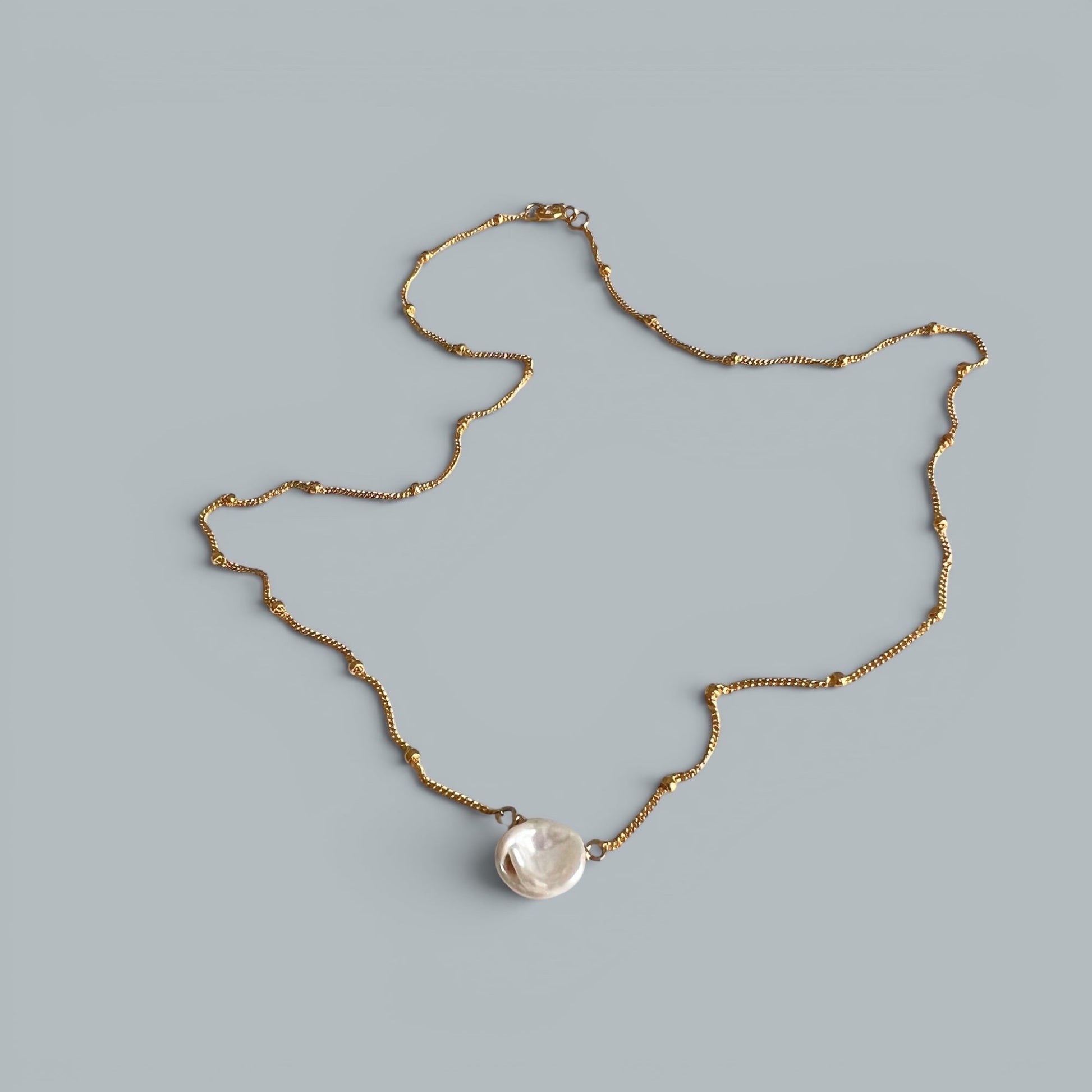 TSJ gold-tone chain necklace with a single, irregular white baroque pearl pendant. Minimalist, elegant design.