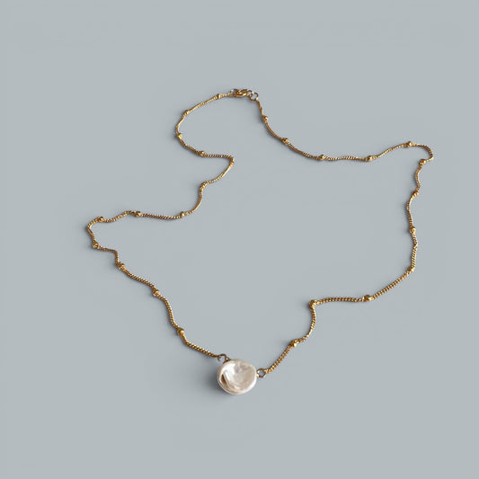 TSJ gold-tone chain necklace with a single, irregular white baroque pearl pendant. Minimalist, elegant design.